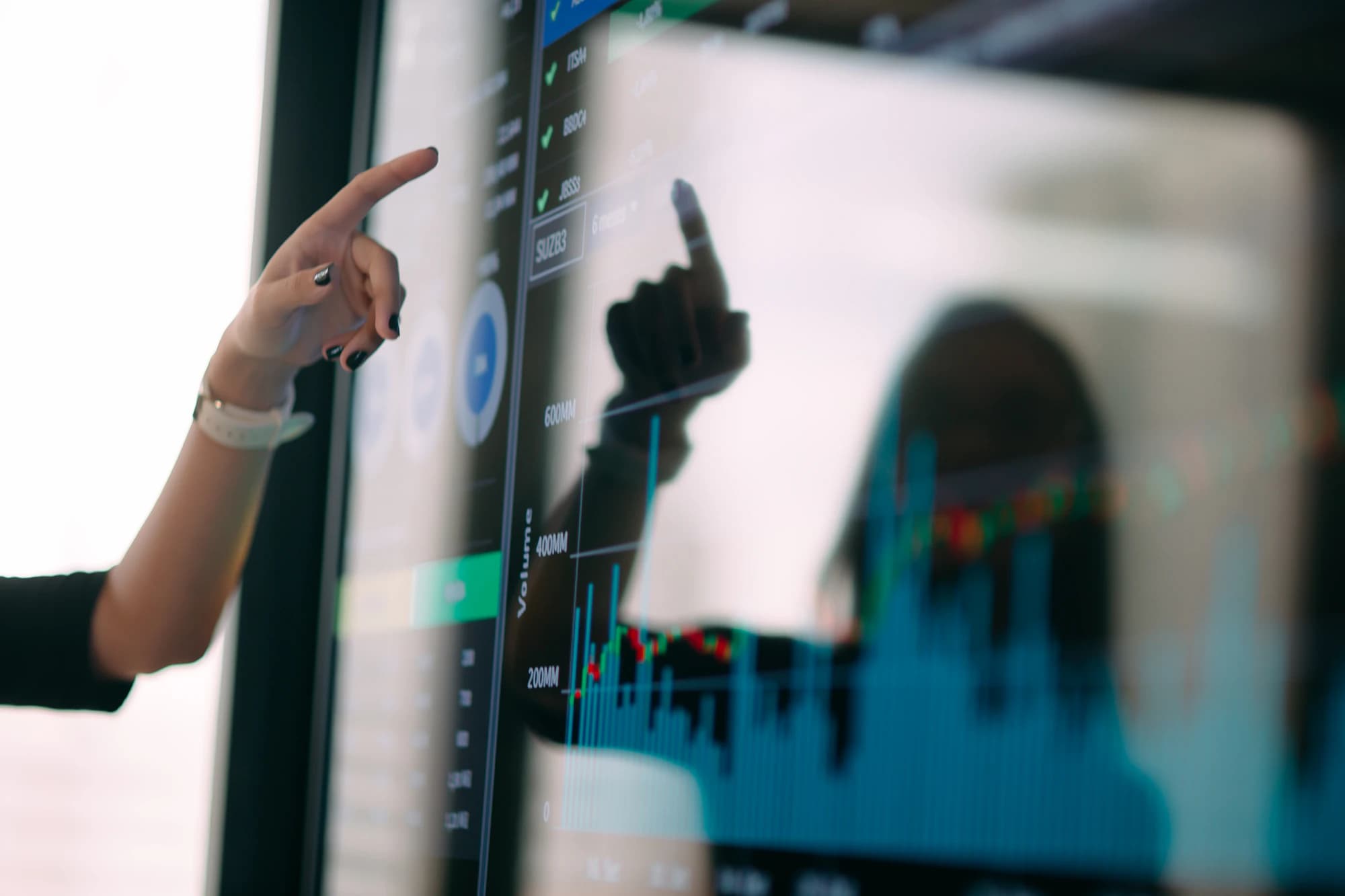 A hand points at a large display of financial charts, with the person's silhouette reflected in the glass screen.