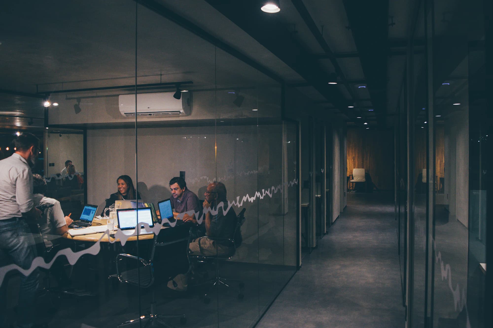 A team meets around a table with laptops inside a glass-walled meeting room off a dim office corridor in the evening.