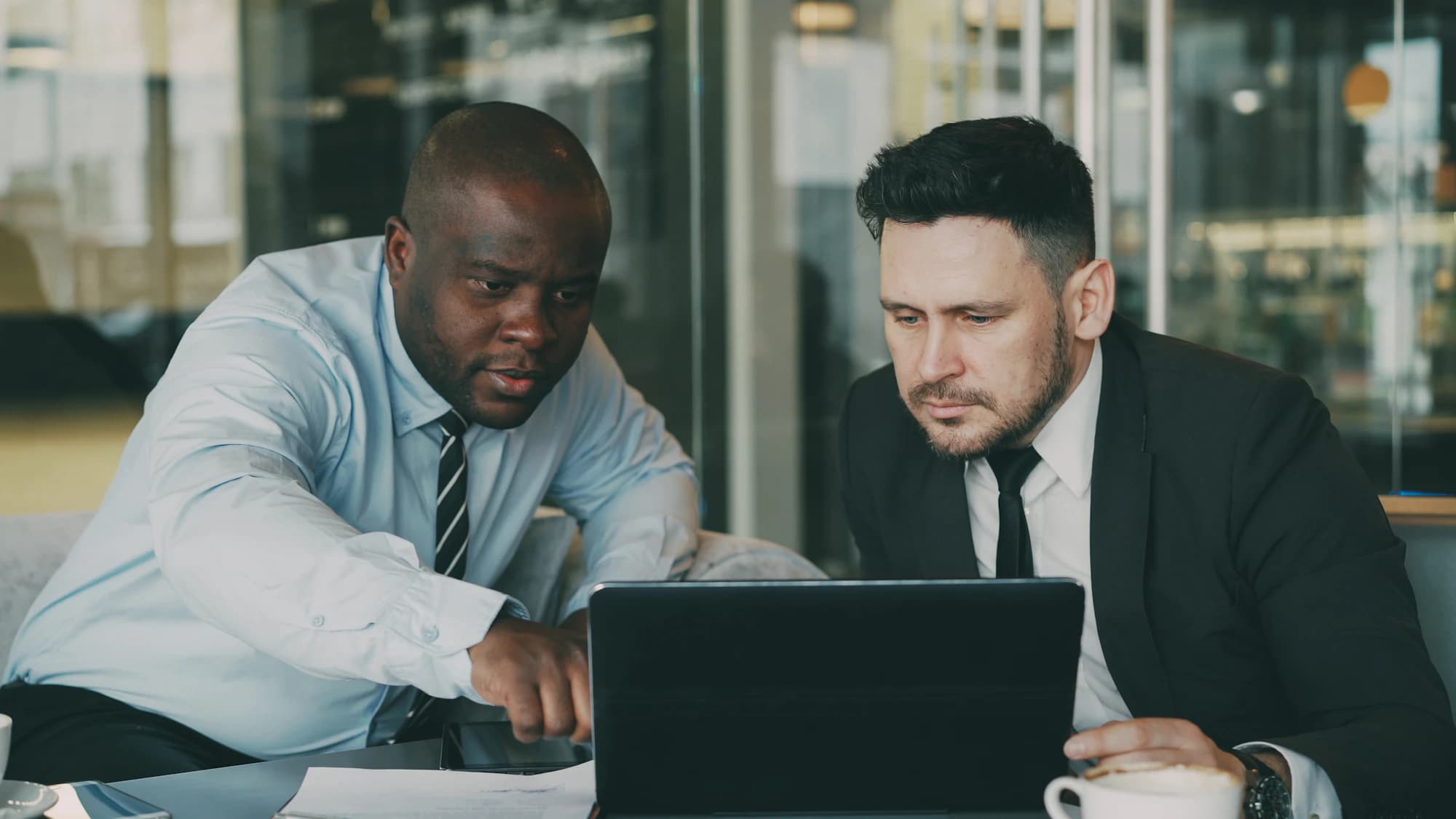 A senior colleague in shirt and tie points at a tablet as he walks a colleague in a dark suit through the work in a glass-walled office.