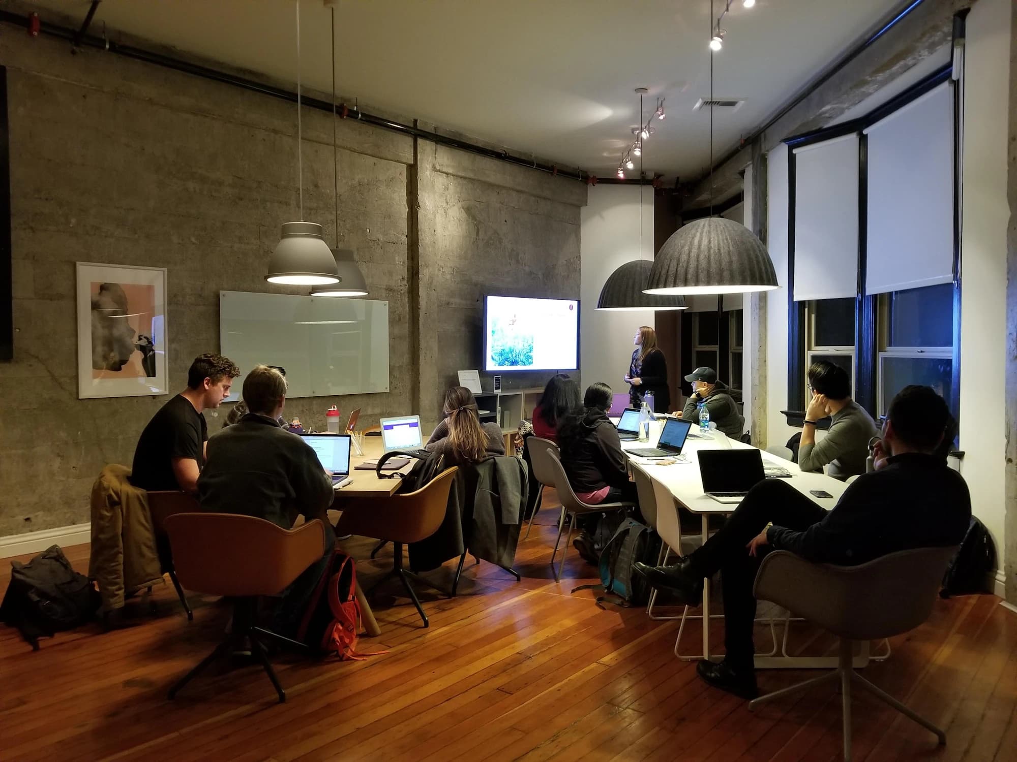 Adults with laptops sit around long tables in a loft-style training room at dusk while a facilitator presents beside a wall screen under pendant lamps.